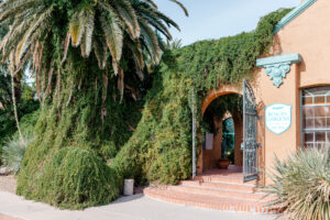 A large, lush palm tree and dense green foliage surround the arched entrance of a stucco building with a turquoise sign and open wrought-iron gate. Sunlight shines on the brick steps and orange exterior walls.