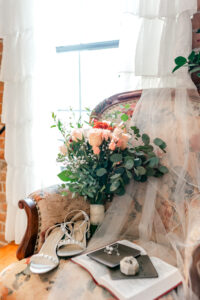 A vintage armchair by a bright window holds a bouquet of flowers, silver sandals, a book, and a ring box with a veil draped over the scene, creating a romantic and elegant atmosphere.