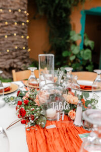 A round table set for an event, decorated with red flowers and greenery, clear glassware, and a clear acrylic sign reading “Table 17” on an orange table runner. A tree wrapped in string lights is in the background.
