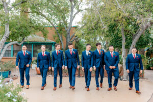 Eight men in matching blue suits, brown shoes, and red ties stand side by side outdoors on a patio, with trees and a building in the background, posing formally for a group photo.