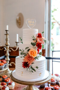 A three-tiered white wedding cake decorated with peach and red flowers and greenery, topped with a heart-shaped “Mr & Mrs” topper, displayed on a gold stand with cupcakes in the background.