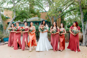 A bride in a white dress stands outdoors with seven bridesmaids in matching rust-colored dresses, all holding bouquets and smiling, surrounded by trees and greenery at Kingan Gardens Tucson Wedding