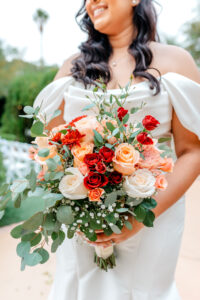 A bride in an off-the-shoulder white dress holds a large bouquet of red, peach, and white roses with greenery and babys breath, standing outdoors with a blurred background.