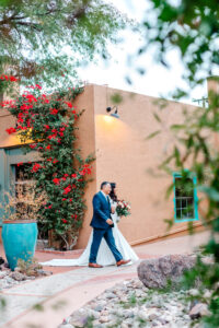 A brides dad walking her outdoors beside an adobe building with red flowers climbing the wall, surrounded by greenery and a turquoise pot.