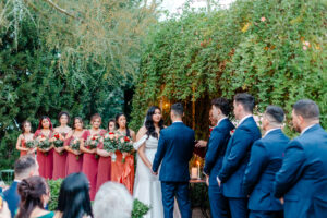 A bride and groom stand facing each other during an outdoor wedding ceremony, surrounded by greenery and guests. The groom is flanked by groomsmen, and tree lights glow warmly in the foreground.