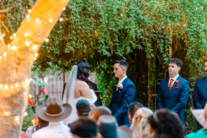 A bride and groom stand facing each other at an outdoor wedding ceremony, surrounded by lush greenery. Bridesmaids in red and pink dresses hold bouquets, while groomsmen in blue suits stand nearby. Guests are seated in the foreground.