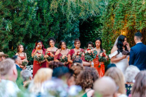 A wedding ceremony outdoors with bridesmaids in matching red dresses holding bouquets, standing beside the bride and groom. Guests are seated in the foreground, surrounded by lush green foliage.