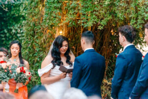 A bride in a white dress reads vows from a book to the groom, who faces her in a navy suit. Bridesmaids with bouquets and groomsmen stand nearby under greenery at an outdoor wedding ceremony.