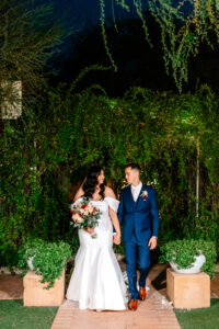 A bride in a white dress and a groom in a blue suit smile at each other while holding hands, standing outdoors at night against a backdrop of lush greenery during Kingan Gardens Tucson Wedding