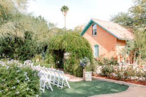 Outdoor wedding setup with rows of white chairs facing a small brick house, surrounded by lush greenery and blooming flowers under a clear sky.
