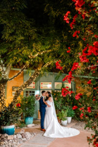A bride and groom share a kiss at night beneath trees adorned with string lights and vibrant red flowers, standing in front of a charming, green-doored building with potted plants around them.