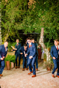 A group of men in blue suits and brown shoes pose playfully outdoors under trees decorated with string lights; one man wears sunglasses and holds a noise maker.