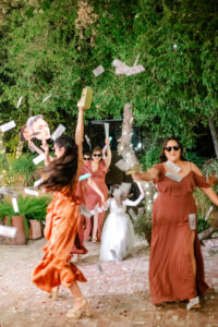 A group of women in festive dresses and sunglasses throw money in the air, celebrating outdoors at night with trees in the background. One woman holds a fan, and a child in white stands behind them.