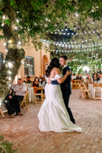 A bride and groom share their first dance outdoors under string lights, surrounded by seated guests at nighttime. The bride wears a white gown, and the groom wears a dark suit. Trees and warm lighting create a romantic atmosphere. at Kingan gardens Tucson Wedding