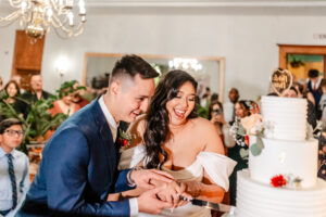 A joyful bride and groom, dressed in formal wedding attire, laugh together as they cut a white tiered wedding cake at their reception, surrounded by smiling guests.