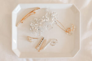 Flat lay of bridal hair accessories including a large silver floral crystal hair comb, gold barrettes, and delicate pearl bobby pins arranged on a white octagonal ceramic tray.
