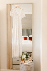 A white lace wedding dress hangs on a "Bride" embossed wooden hanger over a long mirror. At the base of the mirror, white bridal heels, a floral bouquet, and a wooden ring box sit on a grey ottoman.