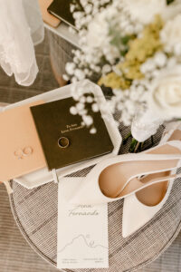 High-angle close-up of wedding details featuring two vow books labeled "To Ana" and "To Fernando," gold wedding bands, white pointed-toe heels, and a wedding invitation with a desert mountain illustration.
