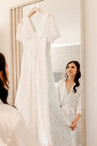A bride in a white silk robe smiles as she looks at her lace wedding dress hanging on a mirror while getting ready for her ceremony in a westin la paloma hotel room