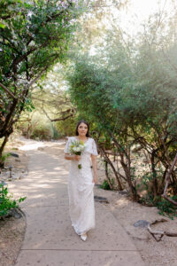 A bride in a short-sleeved beaded lace gown carries a white and yellow bouquet while walking down a sun-drenched desert path at the Westin La Paloma.