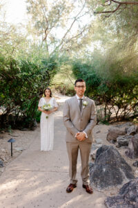 A groom in a tan suit stands on a stone-lined path while his bride approaches him from behind for their "first look" at Westin La Paloma.