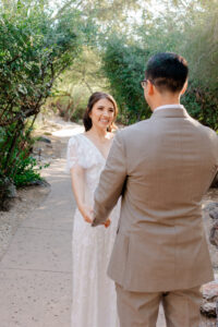 A smiling bride looks up at her groom while holding his hands during their first look on a paved garden path at Westin La Paloma.