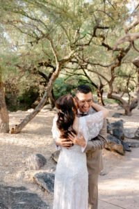 A joyful groom laughs as he pulls the bride into a warm embrace amidst the desert trees and rocky landscape of Westin La Paloma.