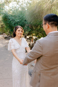 Medium shot of a radiant bride smiling at her groom while holding his hands, featuring the lush green desert foliage of Westin La Paloma in the background.