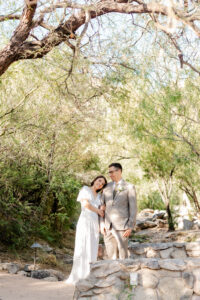 The bride leans her head on the groom's shoulder as they pose together behind a stone wall under a canopy of desert trees at Westin La Paloma.