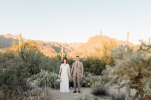 A wide-angle landscape shot of the couple standing on a desert trail in Sabino Canyon, surrounded by saguaros and rocky terrain.