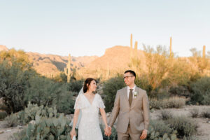 A horizontal landscape shot of the couple standing on a desert path in Sabino Canyon, with the bride leaning her head on the groom's shoulder as they look toward the camera.
