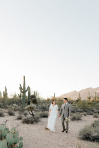 A vertical portrait of the bride and groom standing together on a rocky desert trail, with a tall saguaro cactus prominently featured in the background.