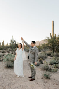 A romantic portrait of the bride and groom facing one another and holding hands on a path lined with desert brush and cacti.