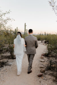 The bride and groom walk hand-in-hand away from the camera down a desert trail in Sabino Canyon