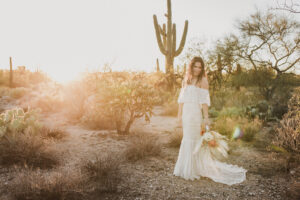 bride holding boho flowers in a boho wedding dress in the desert with cactus behind