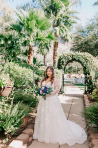Bride in long-sleeve lace ball gown holding navy and burgundy bouquet standing on garden path with palm trees and floral arch at Buttes at Reflections Tucson Arizona