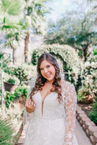 Bride in long-sleeve lace wedding gown smiling and holding her veil in the garden courtyard at Buttes at Reflections wedding venue Tucson Arizona