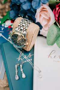 Wedding day flat lay with groom's watch, crystal drop earrings, teardrop pendant necklace, and his and her vow books with blue and blush florals, Tucson wedding photographer
