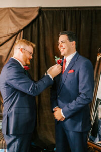 Groomsman pinning a red rose boutonniere on the laughing groom in a navy suit, getting ready at Buttes at Reflections wedding venue Tucson Arizona