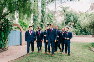 Groom and groomsmen in navy suits with red ties and rose boutonnieres walking and laughing in the garden at Buttes at Reflections Tucson Arizona wedding venue