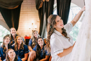 Bride in white satin pajamas revealing her wedding dress to bridesmaids in matching navy pajamas inside the bridal suite at Buttes at Reflections Tucson Arizona