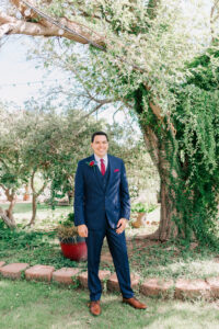 Groom in navy three-piece suit with red tie and rose boutonniere standing in the garden at Buttes at Reflections wedding venue Tucson Arizona