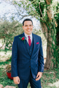 Close-up portrait of groom in navy three-piece suit with burgundy tie and red rose boutonniere in the garden at Buttes at Reflections Tucson Arizona wedding venue