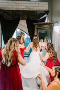 Bridesmaids in red gowns helping bride into lace wedding gown in front of ornate gold mirror inside Buttes at Reflections bridal suite Tucson Arizona