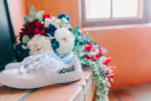 White bridal sneakers reading "Bride" next to a red, white, and navy wedding bouquet