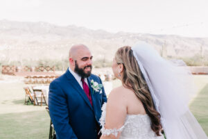 Groom looking at the bride, who is smiling and looking down, with string lights and the venue building behind them