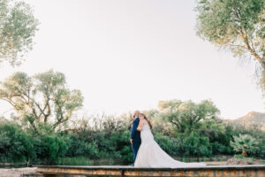 Bride and groom standing together on a wooden dock overlooking a pond lined with cottonwood trees