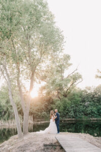 Bride and groom embracing at sunset by the pond, sunlight filtering through the trees
